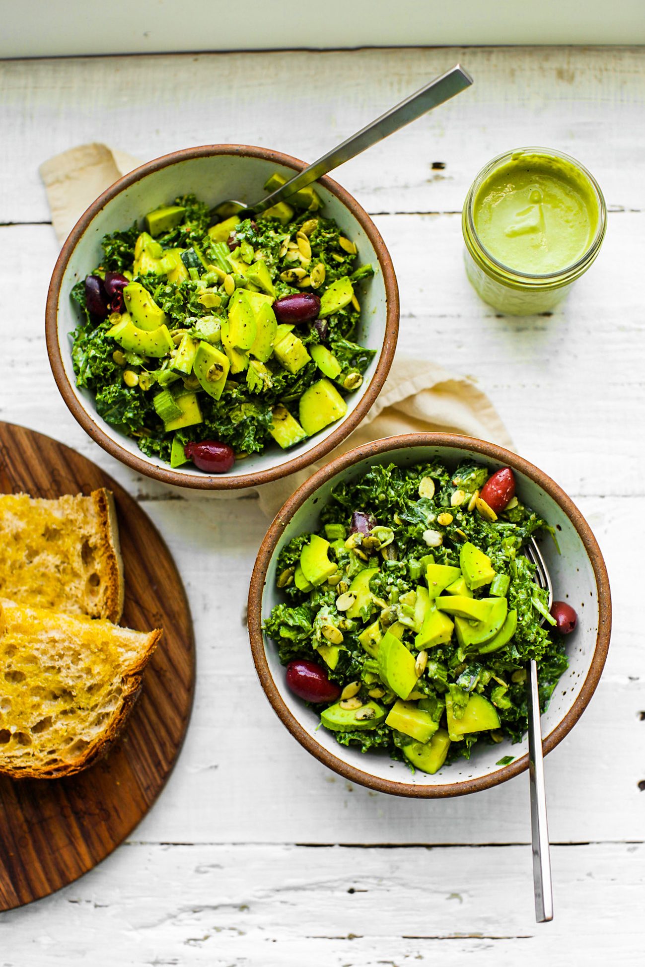 Two bowls of kale-avocado salad with green dressing and crusty bread